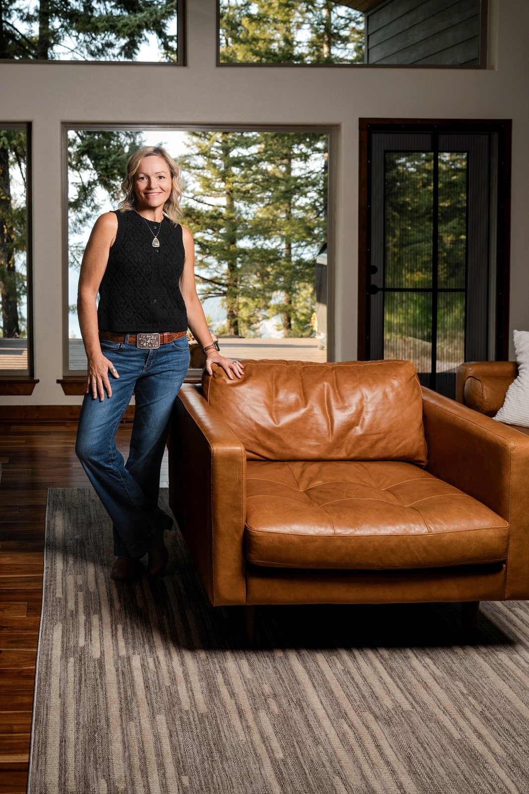 Asenath "CC" Horton, seated in a mountain living room with pine trees and water visible through the windows behind her.